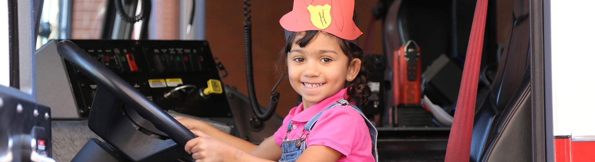 Little girl wears paper fire fighter helmet while sitting in fire truck